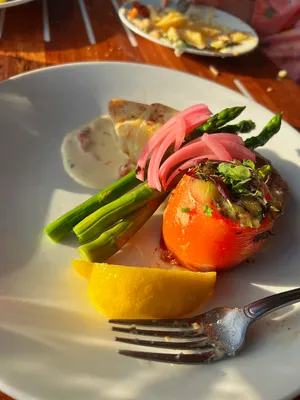 Meal plating at a fancy restaurant with the fork, a lemon wedge, a stuffed tomato, asparagus, pink pickled onion slices, and some fish in a sauce