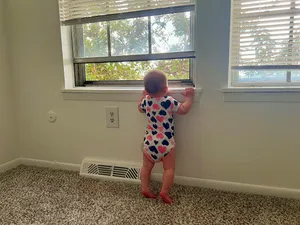 A baby in a heart-pattern onesie pulling up on a window sill to look outside