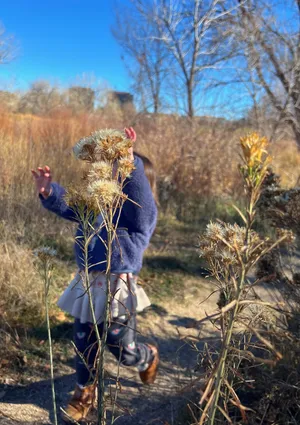 Toddler running in a wild grassy area with arms up like a monster and face obscured by a tall flowering plant